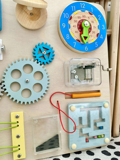 Close-up of wooden Montessori Busy Board with clock, gears, spring lock, maze, and musical elements for open-ended play.